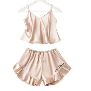 May include: A blush-colored satin pajama set, consisting of a camisole top and shorts, hangs on a white hanger. The top has thin straps and a V-neck. The shorts feature a drawstring waist and a ruffled hem. The words "Mrs. Tribeloy" are printed on the shorts.