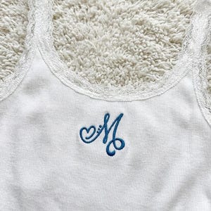 May include: White ribbed tank top with a delicate lace trim along the neckline and straps. The tank top features a blue embroidered letter "M" with heart details. A casual and feminine garment.