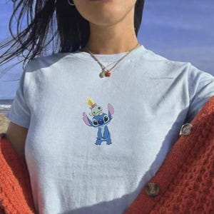 May include: Light blue t-shirt featuring a cartoon image of Stitch, a blue alien character, with a smaller alien on his head. The shirt is made of a soft material and is suitable for casual wear.