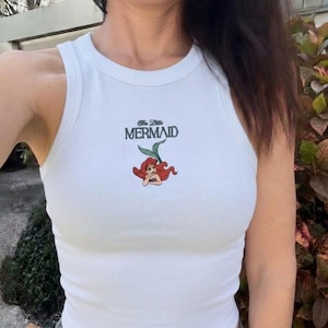 May include: White ribbed tank top featuring the text "The Little Mermaid" in teal and a cartoon illustration of Ariel with red hair and a green tail. The top has a round neckline and a sleeveless design.