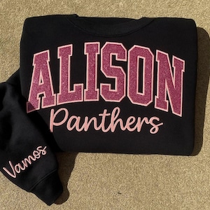 Op de afbeelding: Zwart sweatshirt met de naam "Alison" in glinsterende roze blokletters boven het woord "Panthers" in een scriptlettertype. De linkermouw heeft het woord "Vamos" in een bijpassend roze script. Een casual, sportief kledingstuk.
