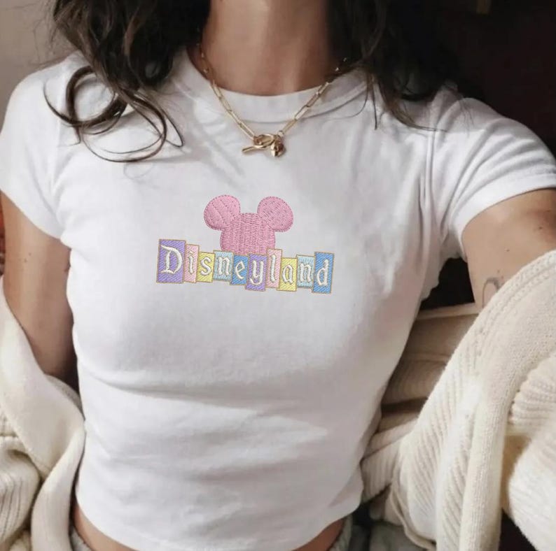 May include: White crop top with a pink embroidered Mickey Mouse head and the word "Disneyland" in pastel colors. The shirt has short sleeves and a crew neck. A gold necklace is visible.