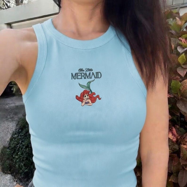 May include: Light blue ribbed tank top featuring the text "The Little Mermaid" above a cartoon illustration of Ariel. The sleeveless top has a scoop neck and is made of a soft, comfortable material.