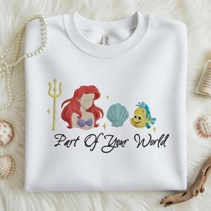 Puede incluir: Sudadera blanca de cuello redondo con un dise&ntilde;o bordado que presenta una sirena pelirroja, un tridente dorado, una concha azul y un pez amarillo. El texto "Part Of Your World" est&aacute; debajo.