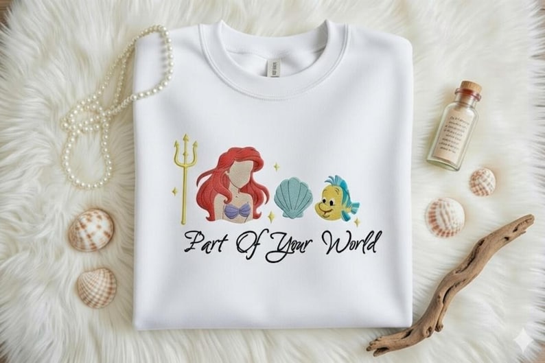 Puede incluir: Sudadera blanca de cuello redondo con un dise&ntilde;o bordado que presenta una sirena pelirroja, un tridente dorado, una concha azul y un pez amarillo. El texto "Part Of Your World" est&aacute; debajo.