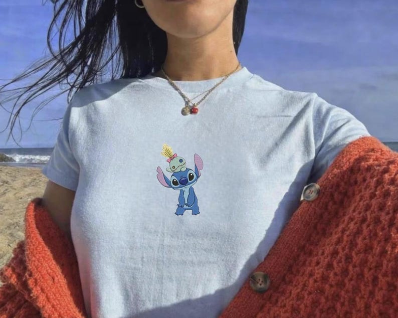 May include: Light blue t-shirt featuring a cartoon image of Stitch, a blue alien character, with a smaller alien on his head. The shirt is made of a soft material and is suitable for casual wear.
