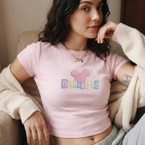 May include: A light pink crop top with short sleeves. The shirt features an embroidered design of a mouse head and the word "Disneyland" in pastel colors. The top is made of a soft material.