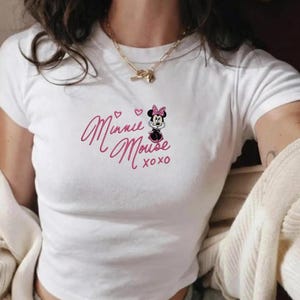Pu&ograve; includere: Top corto bianco con design di Minnie Mouse. Il design include le parole "Minnie Mouse" in rosa, una piccola illustrazione di Minnie Mouse e il testo "xoxo". Il top ha maniche corte.