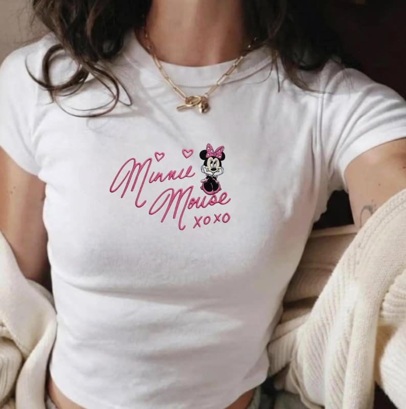 Pu&ograve; includere: Top corto bianco con design di Minnie Mouse. Il design include le parole "Minnie Mouse" in rosa, una piccola illustrazione di Minnie Mouse e il testo "xoxo". Il top ha maniche corte.