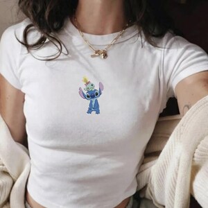 May include: White t-shirt featuring a blue cartoon character with a smaller green character on its head. The graphic is centered on the chest. The shirt has short sleeves and a crew neck.