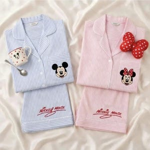 May include: Two sets of Disney-themed pajamas. One set is light blue with Mickey Mouse, the other is pink with Minnie Mouse. Each set includes a button-down shirt and shorts with character embroidery. A Mickey Mouse mug and spoon are next to the blue set.