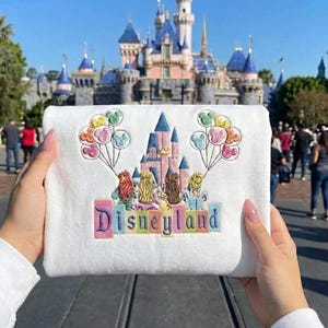 May include: A white sweatshirt with an embroidered design featuring a castle, characters, and Mickey Mouse balloons. The word "Disneyland" is embroidered in colourful letters. The background shows a castle and people.
