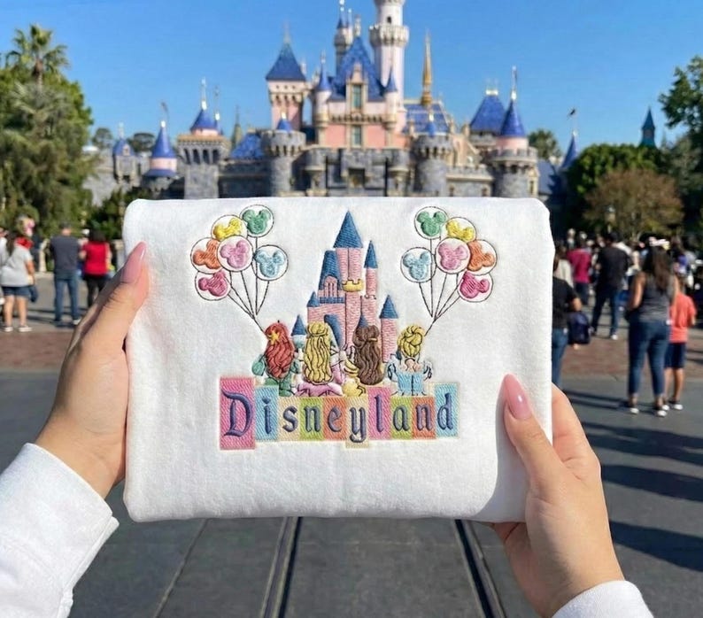 May include: A white sweatshirt with an embroidered design featuring a castle, characters, and Mickey Mouse balloons. The word "Disneyland" is embroidered in colourful letters. The background shows a castle and people.
