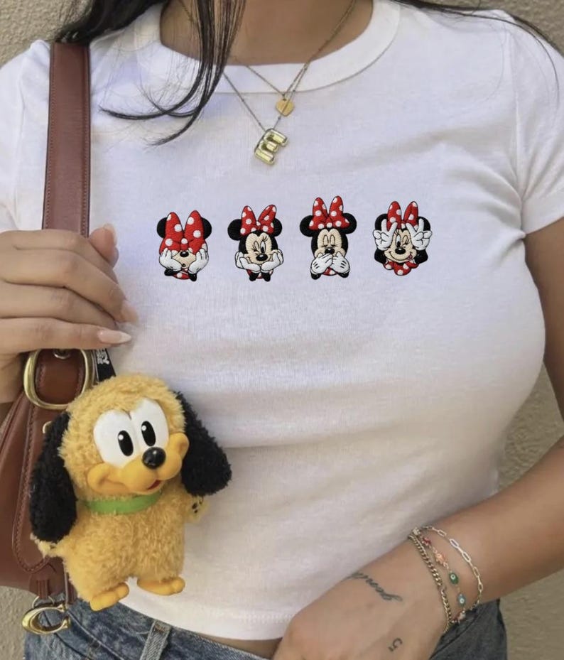 May include: White crew neck t-shirt featuring four Minnie Mouse cartoon graphics with red and white polka dot bows. A yellow plush Pluto keychain hangs from a brown purse strap.