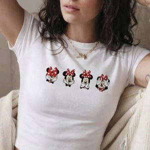 May include: White crop top featuring four Minnie Mouse character designs with red and white polka dot bows. Each Minnie Mouse has a different facial expression. The shirt is a casual, everyday wear item.