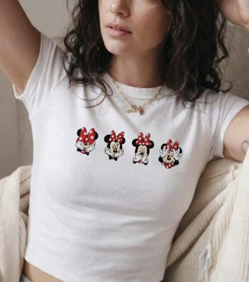 May include: White crop top featuring four Minnie Mouse character designs with red and white polka dot bows. Each Minnie Mouse has a different facial expression. The shirt is a casual, everyday wear item.