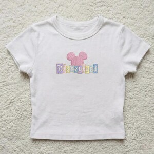 May include: White t-shirt with a pink embroidered Mickey Mouse head and the word "Disneyland" in pastel colors. The shirt has short sleeves and a crew neck, suitable for children.