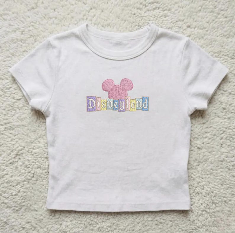 May include: White t-shirt with a pink embroidered Mickey Mouse head and the word "Disneyland" in pastel colors. The shirt has short sleeves and a crew neck, suitable for children.