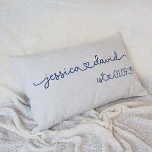 Könnte beinhalten: Ein rechteckiges, hellgraues Kissen mit den Namen "Jessica" und "David" in marineblauer Schrift gestickt, verbunden durch ein Herzsymbol. Darunter ist das Datum "est 01.07.25" ebenfalls in Marineblau gestickt.