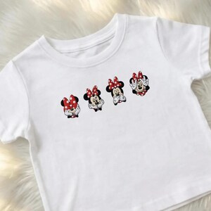 May include: White short-sleeved t-shirt featuring four Minnie Mouse character designs. Each design shows Minnie with a red bow with white polka dots. The cartoon characters are in different poses. The shirt is made of a soft, white fabric.