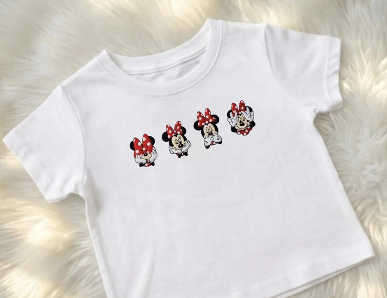 May include: White short-sleeved t-shirt featuring four Minnie Mouse character designs. Each design shows Minnie with a red bow with white polka dots. The cartoon characters are in different poses. The shirt is made of a soft, white fabric.
