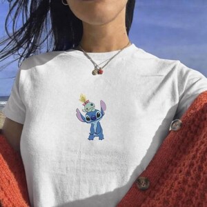 May include: White t-shirt featuring a blue cartoon character with large ears and a smaller green character on top. The graphic is centered on the chest. The person is wearing a rust-colored cardigan.
