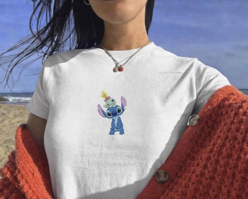 May include: White t-shirt featuring a blue cartoon character with large ears and a smaller green character on top. The graphic is centered on the chest. The person is wearing a rust-colored cardigan.