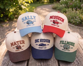 May include: Five baseball caps with off-white crowns and colored brims. Each cap features embroidered text and a character from the Disney Cars franchise: Sally in light blue, McQueen in red, Mater in brown, Doc Hudson in navy, and Fillmore in green.