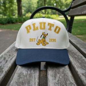 May include: A baseball cap with a white crown and a navy blue brim. The cap features the word "PLUTO" in gold lettering, along with an embroidered image of Pluto and the text "EST 1930". The cap is resting on a wooden bench.