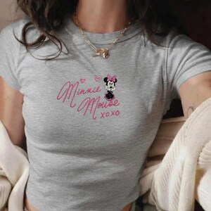Pu&ograve; includere: Un crop top grigio con scollo tondo. La parte superiore presenta un ricamo rosa con la scritta "Minnie Mouse xoxo" e un disegno di Minnie Mouse con fiocco rosa e vestito nero.