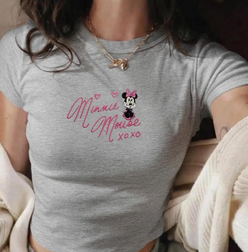 Pu&ograve; includere: Un crop top grigio con scollo tondo. La parte superiore presenta un ricamo rosa con la scritta "Minnie Mouse xoxo" e un disegno di Minnie Mouse con fiocco rosa e vestito nero.