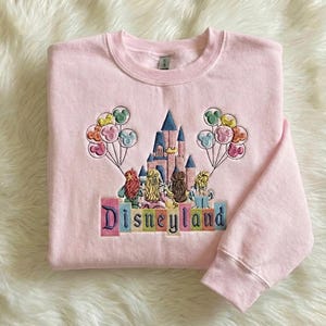 May include: A pale pink sweatshirt featuring an embroidered Disneyland design. The design includes a castle, characters, and balloons. The word "Disneyland" is embroidered in colourful letters. The sweatshirt has a crew neck and long sleeves.