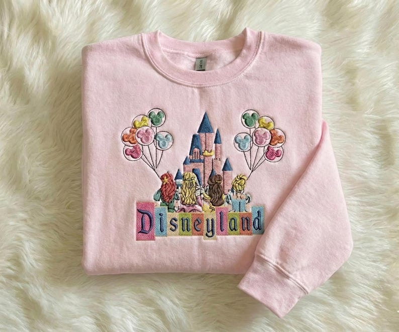 May include: A pale pink sweatshirt featuring an embroidered Disneyland design. The design includes a castle, characters, and balloons. The word "Disneyland" is embroidered in colourful letters. The sweatshirt has a crew neck and long sleeves.