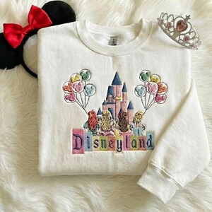 May include: White sweatshirt featuring a Disneyland castle embroidery, balloons, and the word "Disneyland". Includes a red bow Minnie Mouse ear headband and a silver tiara.