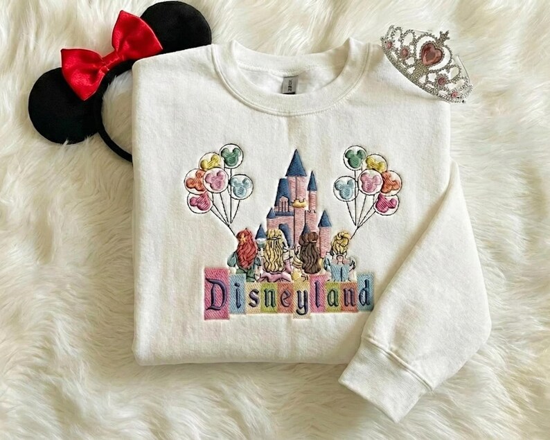 May include: White sweatshirt featuring a Disneyland castle embroidery, balloons, and the word "Disneyland". Includes a red bow Minnie Mouse ear headband and a silver tiara.