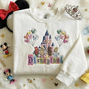 May include: White crew neck sweatshirt featuring an embroidered Disneyland castle design. The design includes colourful balloons, princess figures, and the word "Disneyland" in pastel letters. Accessories include Minnie Mouse ears, figurines, and a tiara.