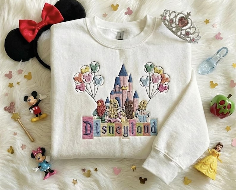 May include: White crew neck sweatshirt featuring an embroidered Disneyland castle design. The design includes colourful balloons, princess figures, and the word "Disneyland" in pastel letters. Accessories include Minnie Mouse ears, figurines, and a tiara.