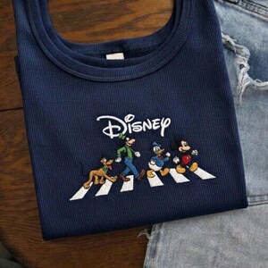 May include: Navy blue ribbed tank top featuring the Disney logo and an embroidered design of Disney characters crossing a white crosswalk. The characters include Goofy, Donald Duck, and Mickey Mouse.