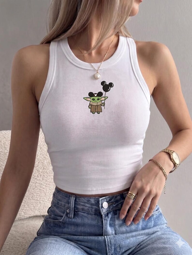 May include: White ribbed tank top featuring a cartoon image of Grogu, also known as Baby Yoda, holding a black Mickey Mouse balloon. The tank top is sleeveless and has a round neckline. The design is centered on the front of the top.