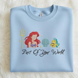 Puede incluir: Sudadera azul claro con un dise&ntilde;o bordado que presenta un tridente dorado, una sirena pelirroja, una concha y un pez amarillo. El texto "Part Of Your World" est&aacute; escrito debajo.