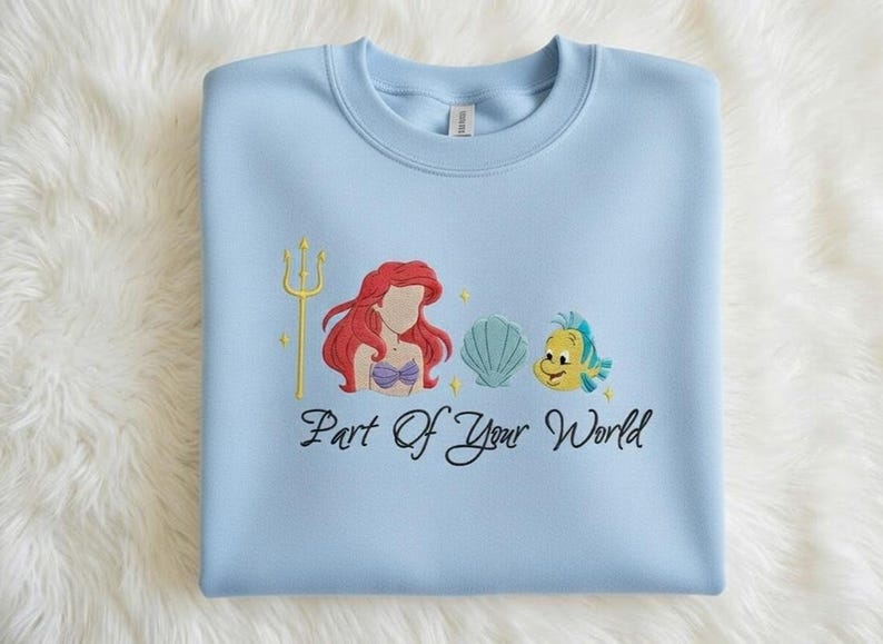 Puede incluir: Sudadera azul claro con un dise&ntilde;o bordado que presenta un tridente dorado, una sirena pelirroja, una concha y un pez amarillo. El texto "Part Of Your World" est&aacute; escrito debajo.