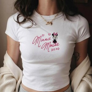 Pu&ograve; includere: T-shirt bianca a maniche corte con un design ricamato rosa. Il design presenta Minnie Mouse e il testo "Minnie Mouse xoxo", insieme a due cuori e un'immagine a cartoni animati di Minnie Mouse con un fiocco rosa.