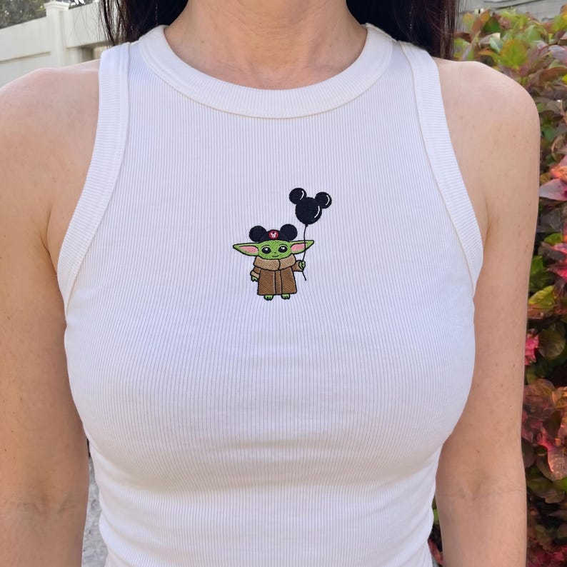 May include: White ribbed tank top featuring an embroidered image of Grogu, also known as Baby Yoda, holding a black Mickey Mouse balloon. The character is wearing a brown coat and a pink bow. The tank top has a round neckline.