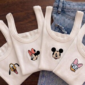 May include: Four white ribbed tank tops with cartoon character designs. Each tank top features a different character: Pluto, Minnie Mouse, Mickey Mouse, and Daisy Duck. The tank tops are displayed on a denim background.