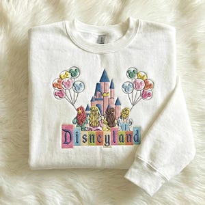 May include: White crew neck sweatshirt with an embroidered design featuring a castle, characters, and balloons. The word "Disneyland" is embroidered in colourful letters across the bottom of the design.