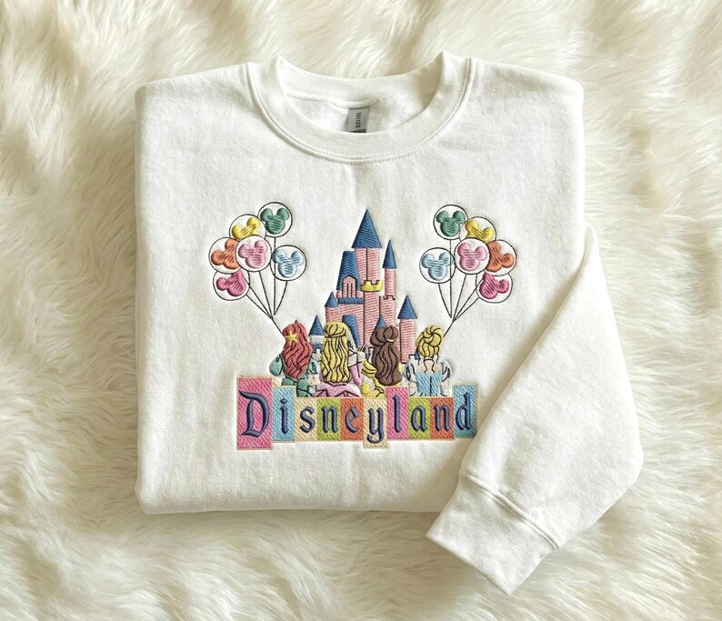 May include: White crew neck sweatshirt with an embroidered design featuring a castle, characters, and balloons. The word "Disneyland" is embroidered in colourful letters across the bottom of the design.