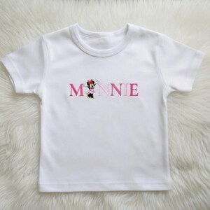 May include: White short-sleeved t-shirt with the name "MINNIE" embroidered in pink letters. The letter "M" features a cartoon character in a red bow. The shirt is made of a soft, white fabric.