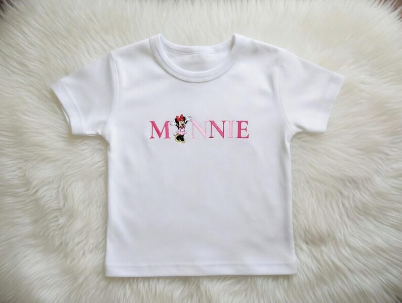 May include: White short-sleeved t-shirt with the name "MINNIE" embroidered in pink letters. The letter "M" features a cartoon character in a red bow. The shirt is made of a soft, white fabric.