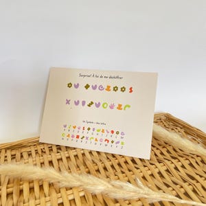 Può includere: Un biglietto di auguri con il testo "Surprise! À toi de me déchiffrer" e una serie di simboli colorati. Sotto, il biglietto mostra un alfabeto con ogni lettera rappresentata da un simbolo unico. Il biglietto è su un cestino intrecciato.