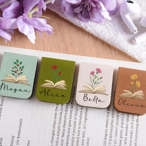 Può includere: Quattro segnalibri in pelle con disegni floreali e nomi: Megan, Alice, Bella e Olivia. Ogni segnalibro presenta un'illustrazione di un libro aperto. I segnalibri sono in tonalità di verde, beige e marrone.